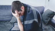 Symbolbild: Ein Mensch sitzt unter eine Decke auf einem Sofa und stützt den Kopf in die Hand.