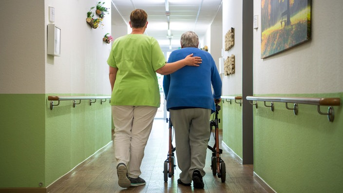 Eine Pflegekraft geht mit einer älteren Frau mit Rollator über einen Flur. Symbolbild 