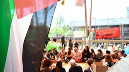 Studierende sitzen unter Plakaten und Palästinaflaggen im Kreis