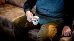Älterer Mann mit Kaffeetasse sitzt auf einem Sofa. Aufnahme vom Hals abwärts.