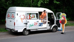 Ein Eisverkäufer mit orangenem T-Shirt lehnt sich aus seinem weißen Eiswagen mit der Aufschrift "Ital. Eis".