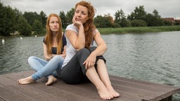 Zwei junge Frauen sitzen auf einem Steg an einem See, ohne sich dabei anzusehen.