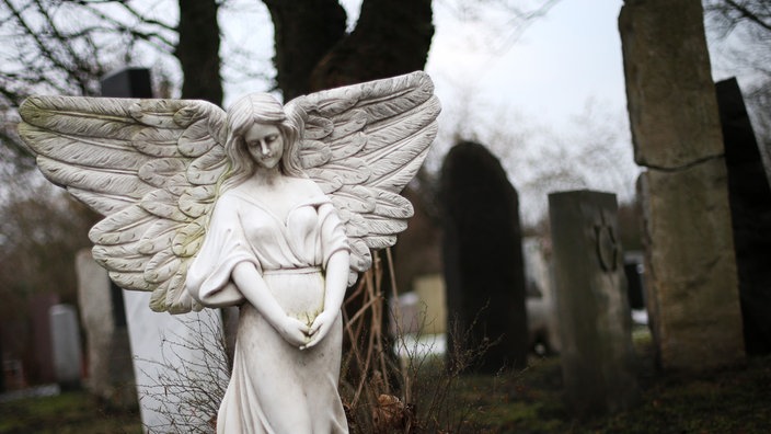 Eine Engelsfigur als Grabstein steht auf einem Friedhof.