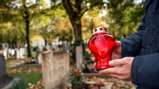 Ein Mann hält bei einem Friedhofsbesuch eine rote brennende Grabkerze in der Hand vor einem Grab. 