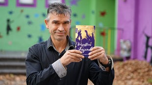 Hermann Wenning mit grauem Haar im Hemd, hält sein Buch in die Kamera.