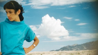 Joan Baez in einem türkis-blauen Oberteil und karierter Hose schauen mit den Händen in die Hüften gestemmt in Richtung Boden, vor blauem Himmel einer Wüstenlandschaft.