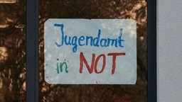 "Jugendamt in Not" steht auf einem Plakat, das an einem Fenster befestigt ist. 