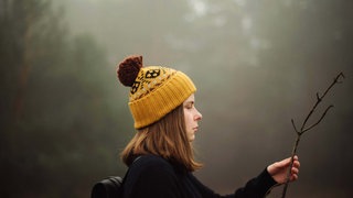 Junge Frau mit Pudelmütze blickt auf einen Ast, den sie in der Hand hält.