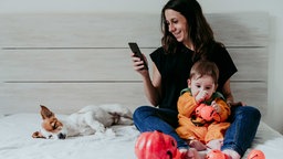 Eine Frau sitzt mit ihrem Kind auf dem Bett und schaut auf ihr Smartphone, neben ihr schläft ein Hund.