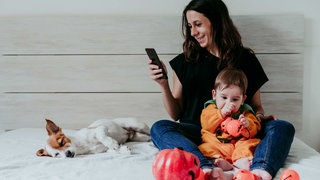 Eine Frau sitzt mit ihrem Kind auf dem Bett und schaut auf ihr Smartphone, neben ihr schläft ein Hund.