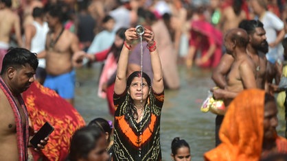 Eine Frau in indischen Gewändern steht im Wasser des für Hindus heiligen Flusses Ganges und schüttet einen Tasse mit dessen Wasser in Kopfhöhe aus.