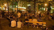 Szenetreff Brüsseler Platz vor der Kirche St. Michael im Sommer. Bild: Menschen stehend, sitzend, erleuchtet