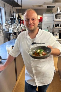 Sternekoch Maximilian Lorenz steht in seiner Küche und hält einen Teller Lachs Tataki