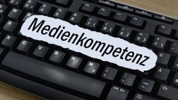 Eine schwarze Computer-Tastatur auf der ein Zettel mit der Aufschrift "Medienkompetenz" liegt.