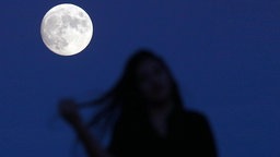 Ein junge Frau steht schemenhaft vorm Vollmond und fährt mit einer Hand durch ihr langes Haar