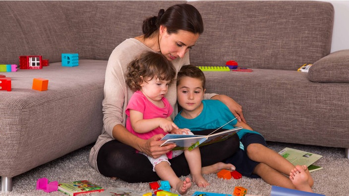 Eine Mutter sitzt umringt von Spielzeug mit zwei Kindern vor einem Sofa und liest ihnen aus einem Buch vor.