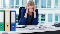 Eine Frau in einem blauen Blazer sitzt verzweifelt, den Kopf auf ihre Häne stützend, in einem hellen Büro.