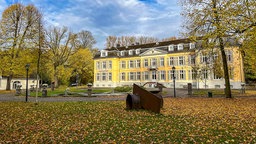 Ein gelb gestrichenes Schloss in einer Gartenanlage mit gepflasteter Einfahrt.