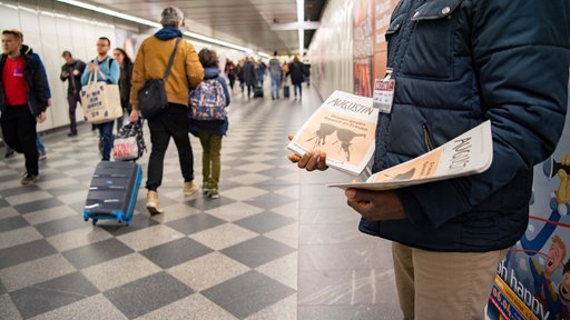 Mann steht in einer U-Bahn-Station und bietet Straßenzeitung zum Verkauf an. 