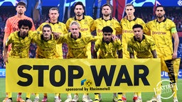 Die Mannschaft von Borussia Dortmund hält ein Banner, darauf steht: Stop War - sponsored by Rheinmetall