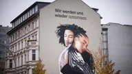 Mural auf Hausfassade: zwei Menschen umarmen einander, darüber der Schriftzug "Wir werden uns wieder umarmen"