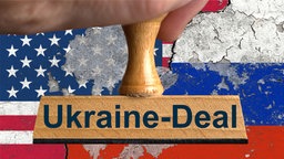 Holzstempel mit Aufschrift "Ukraine-Deal", gehalten vor den Nationalflaggen der USA und der Russischen Föderation im Anschnitt auf einer Wand mit abgeplatzter und rissiger Wandfarbe, 13.02.2025.