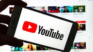 Das Logo des Video-Portals YouTube wird auf dem Display eines Smartphones angezeigt. Im Hintergrund ist auf einem Bildschirm die YouTube Homepage zu sehen