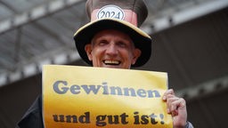 DFB-Fußball-Fan hält lachend Schild mit Aufschrift "Gewinnen und gut ist" in der Hand. 