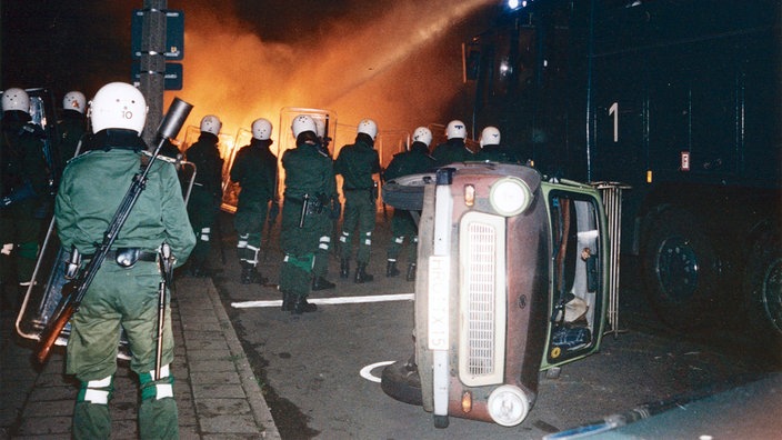 Polizeibeamte bekämpfen am Abend des 24. August 1992 in Rostock-Lichtenhagen eine brennende Strassenblockade mit einem Wasserwerfer. Im Vordergrund liegt ein Auto auf der Seite