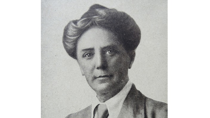 Dame Ethel Mary Smyth schaut hintergründig fein lächelnd in die Kamera, sie trägt einen Anzug mit Hemd und Krawatte, ihr schlankes Gesicht wird von einer Hochsteckfrisur umrahmt