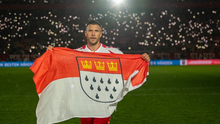 Lukas Podolski steht mit einer großen Köln-Fahne im RheinEnergieSTADION