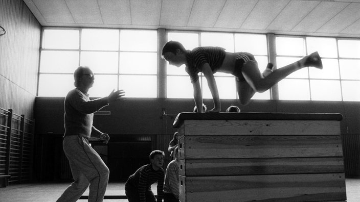 Ein Sportlehrer gibt einem Schüler Hilfestellung in der Turnhalle