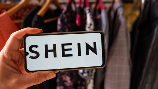 Das Bild zeigt eine Kleiderstange und im Vordergrund hält eine Hand ein Mobiltelefon, auf dem das Shein-Logo zu sehen ist. 