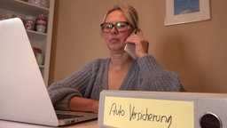 Eine Frau telefoniert mit dem Handy. Im Vordergrund ein Ordner mit der Aufschrift "Auto Versicherung"