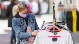 Das Bild zeigt eine Frau mit Maske, die sich Kleidung auf einer Kleiderstange ansieht.