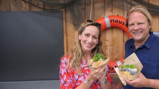 Tamina Kallert und Björn Freitag stehen auf einem Steg im Hafen Lübecks und haben Fischbrötchen in der Hand.