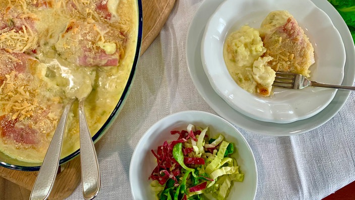 Fenchelgratin in einer Glasform sowie eine Portion auf einem Teller, daneben eine Schüssel mit Salat