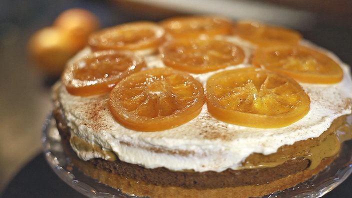 Auf einer geschichteten Torte aus dunklem Teig liegen glänzende Orangen-Scheiben auf einer weißen Creme.