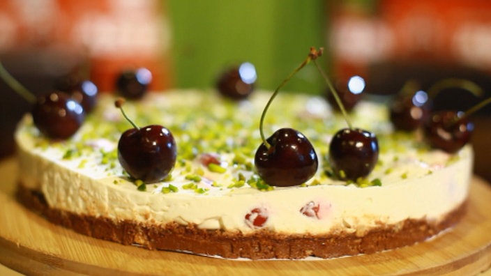 Ein Kuchen mit dunklem Boden, heller Frischkäsecreme ist mit Pistazien und Kirschen bedeckt.