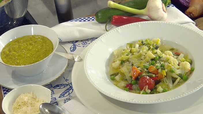 Minestrone in einem Teller angerichtet, daneben Schüsseln mit Basilikumsauce und geriebenem Parmesan