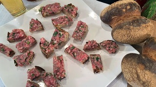 Röggelchen-Happen mit Mett auf einer Platte, daneben ein Glas Bier und ganze Röggelchen