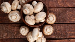 Einige Champignons liegen in einer Holzschale und auf einem Holztisch.