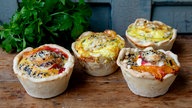 Das Bild zeigt Mini-Quiches aus einer Muffinform auf einem Holzbrett.