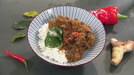 Das indonesische Gericht "Beef Rendang" in einer tiefen Schüssel, drumherum liegen Chilischoten und Ingwer.