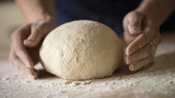 Zwei Hände formen einen Brotteig auf einer mit Mehl bestreubten Arbeitsfläche.